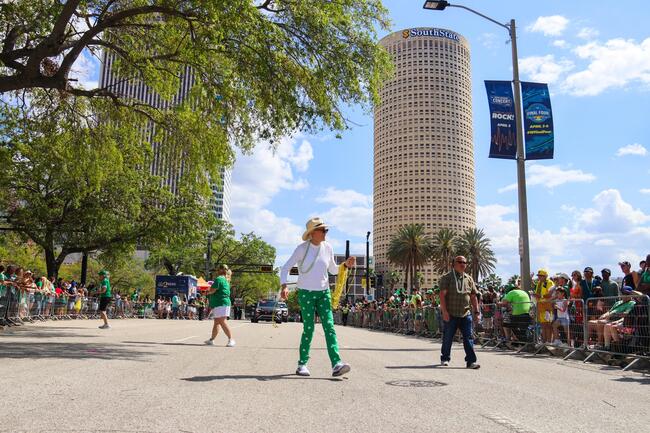 Parade with people in festive outfits on a sunny street, tall buildings in the background.