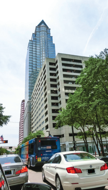 City street with cars and a blue bus, tall multi-story parking garage in the background, trees lining the road.