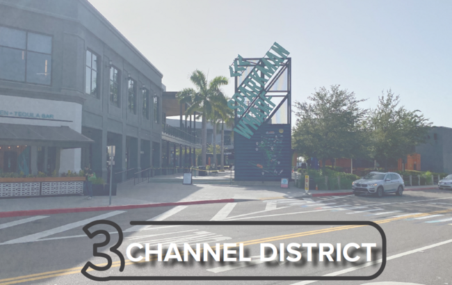 Channel District street scene with palm trees and modern buildings in sunlight.