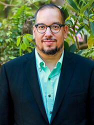 Man in glasses and suit jacket, standing outdoors with greenery in the background.
