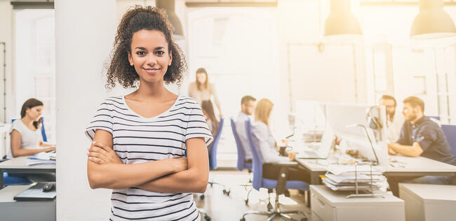 Woman in a striped shirt, arms crossed, standing in a bright office space.
