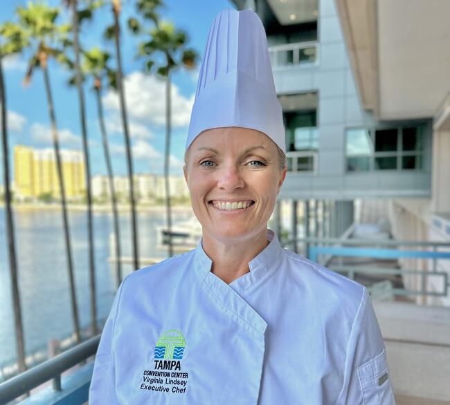 A professional portrait of Executive Chef Virginia Ginny Lindsey at the Tampa Convention Center, wearing a white chef jacket and tall toque. The TCC logo and her title are embroidered on her jacket. The background features the TCC exterior, palm trees, and the waterfront under a bright blue sky.