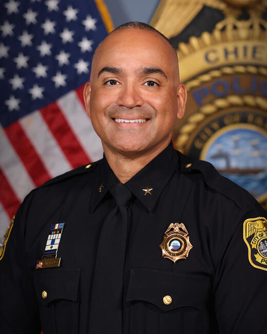 Photograph of a smiling police chief in uniform, with a US flag and police badge backdrop, June 2026.