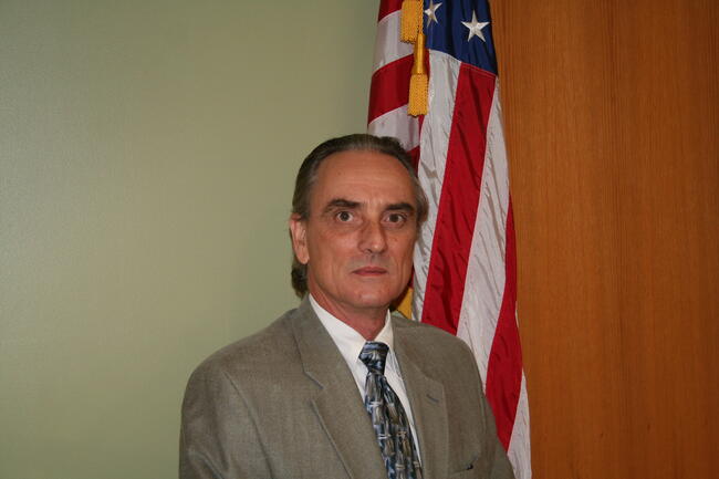 Commissioner Stephen Sutton. A man in a suit standing in front of a U.S. flag and a wood-paneled wall.