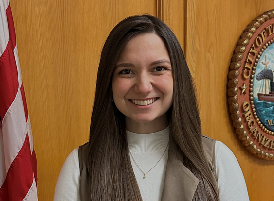 Photo of Comissioner Ansley Wright smiling, standing in front of a wooden wall and official seal.