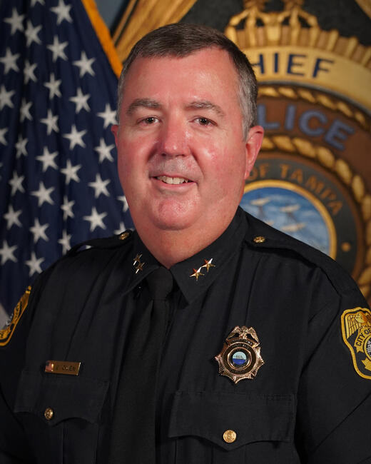 Photograph of Assistant Chief Brett Owen in police uniform, 2025, with badge and flag in background.