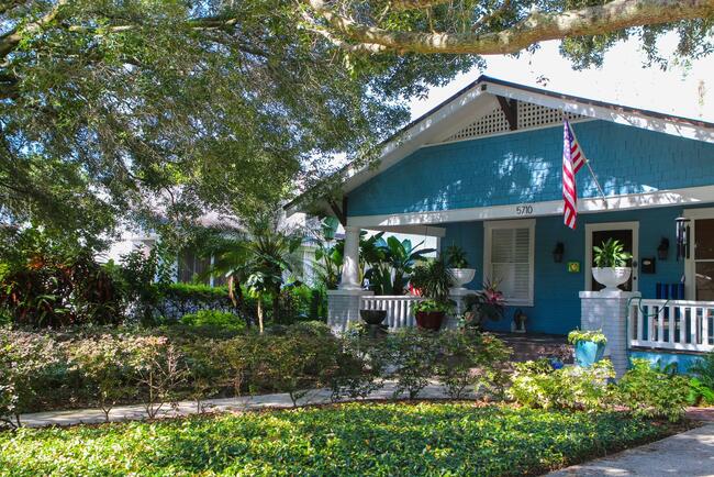 Blue house with front porch, garden, and an American flag under large shade trees.