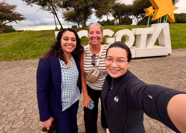 Three people smiling in front of large white letters spelling "Bogotá" outdoors.