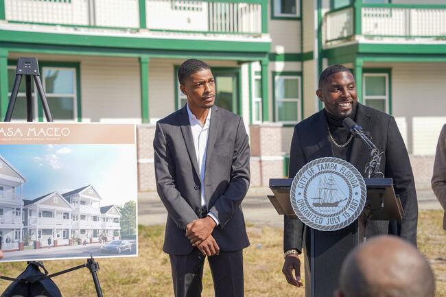 Two men speak at a Casa Maceo press conference outdoors, with building plans displayed nearby.