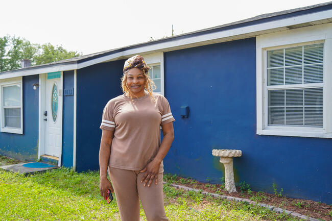 Smiling woman standing in front of a blue house with white trim and a small garden bench.