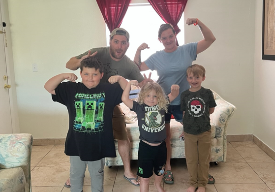 Smiling family of five posing and flexing arms in a cozy, sunlit living room.