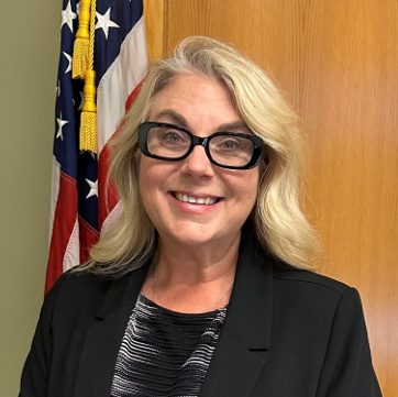 Professional photo of Commissioner Dr. Commissioner Monika Alesnik smiling, standing by a U.S. flag and wooden wall.