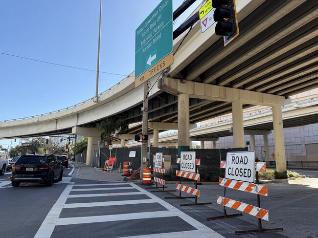 Road closed signs and barricades block a city street under a highway overpass.