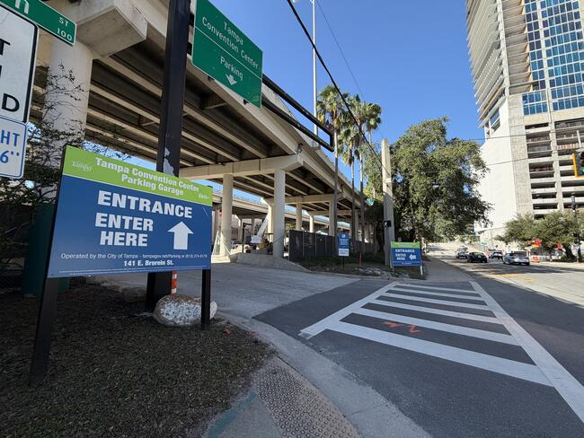 Blue entrance signs by a sidewalk under a highway overpass in a city setting.