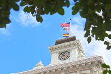 Clock tower with waving flags against a blue sky, framed by tree leaves.