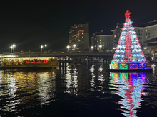 Lit holiday tree on river with city skyline; a festive boat floats nearby.