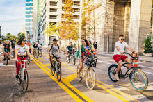 People cycling on city street with tall buildings and trees in background.