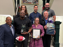 Group photo with award recipients holding a trophy, certificate, and gift basket indoors.