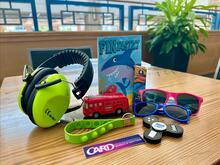 Colorful items on a table: headphones, toy bus, sunglasses, wristbands, fidget spinner, "Pinwheel" book.