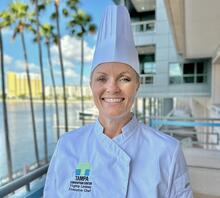 A professional portrait of Executive Chef Virginia Ginny Lindsey at the Tampa Convention Center, wearing a white chef jacket and tall toque. The TCC logo and her title are embroidered on her jacket. The background features the TCC exterior, palm trees, and the waterfront under a bright blue sky.