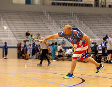 Man in Captain America shirt playing badminton in a large indoor sports arena.