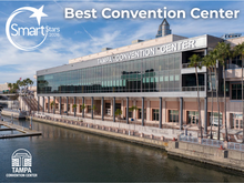 An award announcement graphic featuring an elevated, daytime view of the Tampa Convention Center’s waterfront exterior. The multi-story concrete and glass building stretches along the water, flanked by palm trees and empty boat slips. In the top left corner is a white "Smart Stars 2026" logo featuring a star crescent design, next to large white bold text reading "Best Convention Center." The white line-art logo for the Tampa Convention Center is positioned in the bottom left corner against the water.