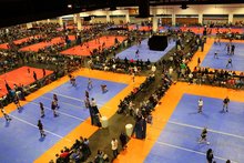 Arial view of the Tampa Convention Center exhibit hall shows blue and orange volleyball courts set up and thousands of athletes, coaches, and parents spectating the tournament.