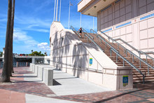 Photo shows the new ADA ramp on the Tampa Riverwalk. To the right of the ramp is the staircase leading up to the flag deck. The Riverwalk sidewalk is to the left of the ramp. The sky is blue in the background.
