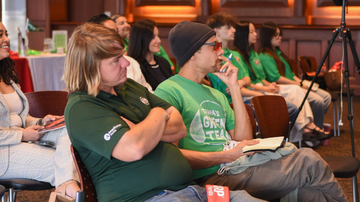 People in matching green shirts attentively watch a presentation indoors at Heat 2 event.