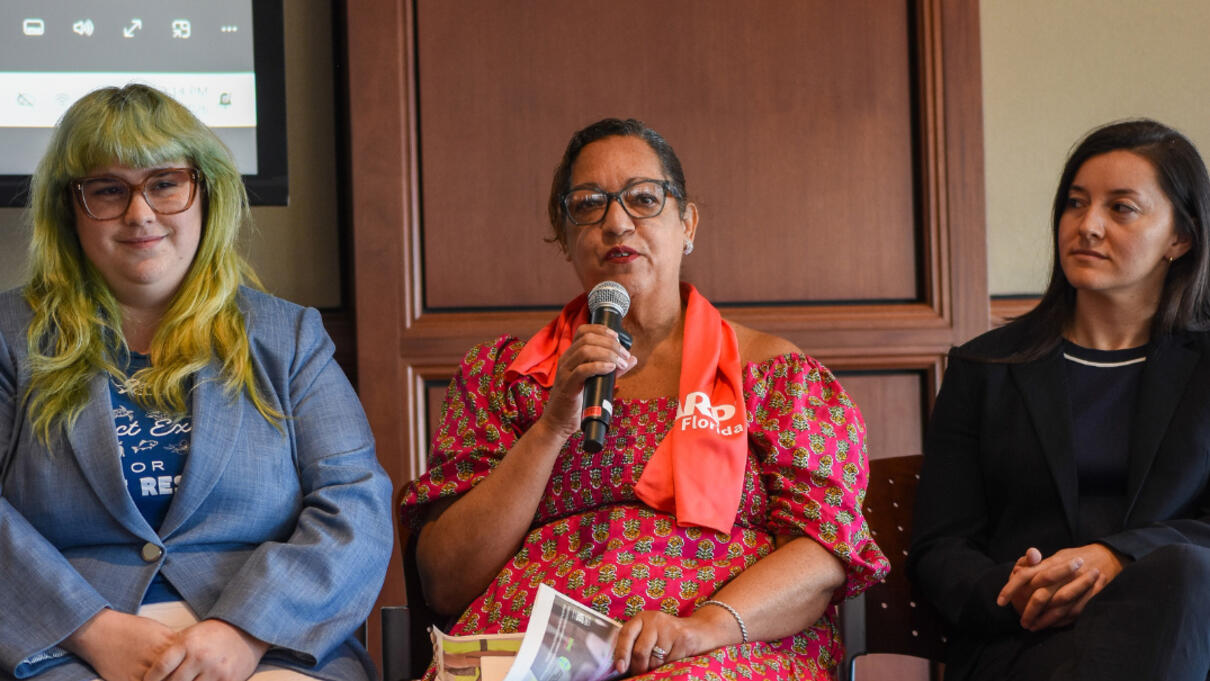 Three women seated indoors, one speaking into a microphone during a panel discussion.