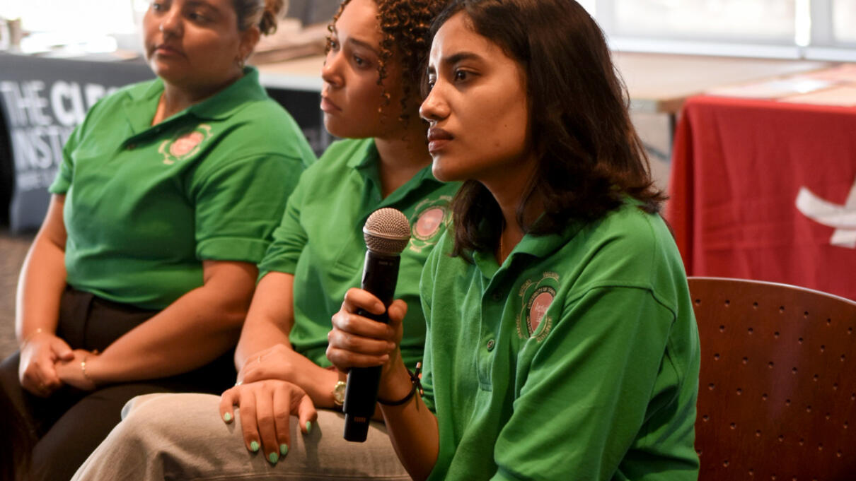 Three women in green shirts sit indoors, one holding a microphone, during a heat discussion panel.