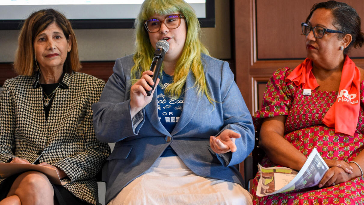 Three women sit on a panel, one speaking into a microphone, in a discussion setting.