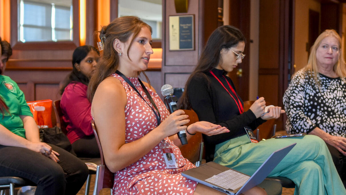 Woman with a microphone speaks during a discussion in a conference room, others listen nearby.