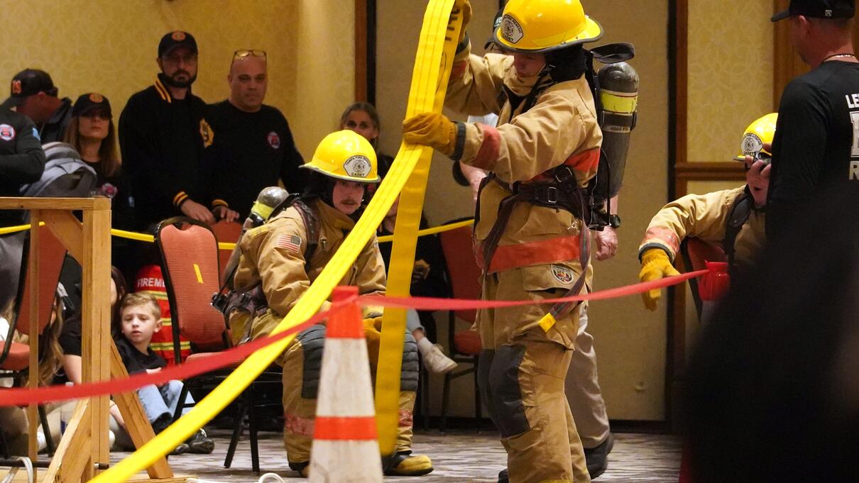 Firefighters in gear participating in an indoor fire safety competition event with onlookers.