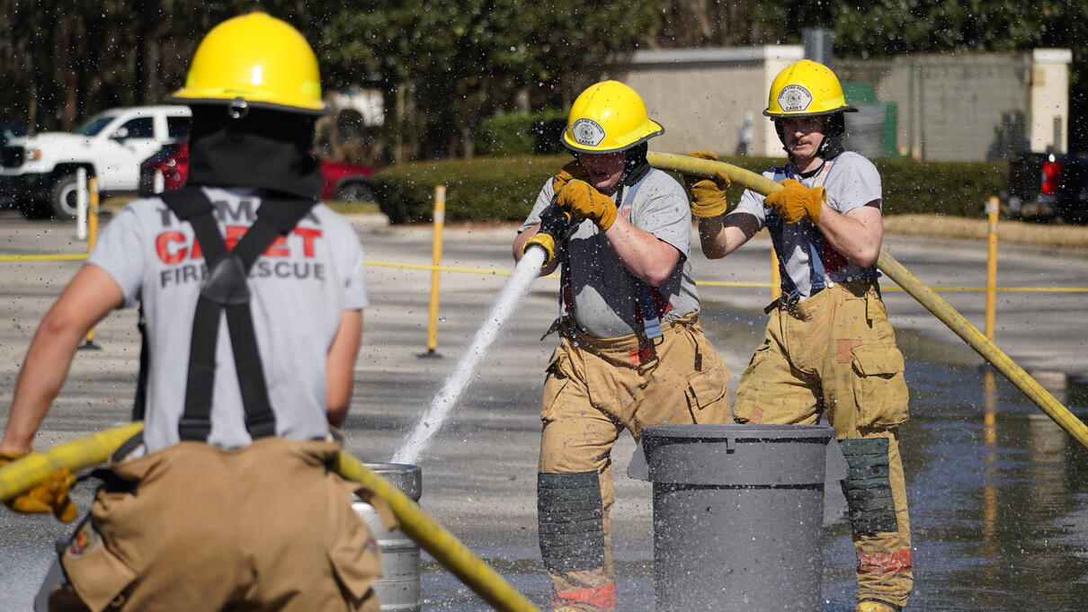 Firefighter trainees in yellow helmets using hoses in an outdoor training exercise.