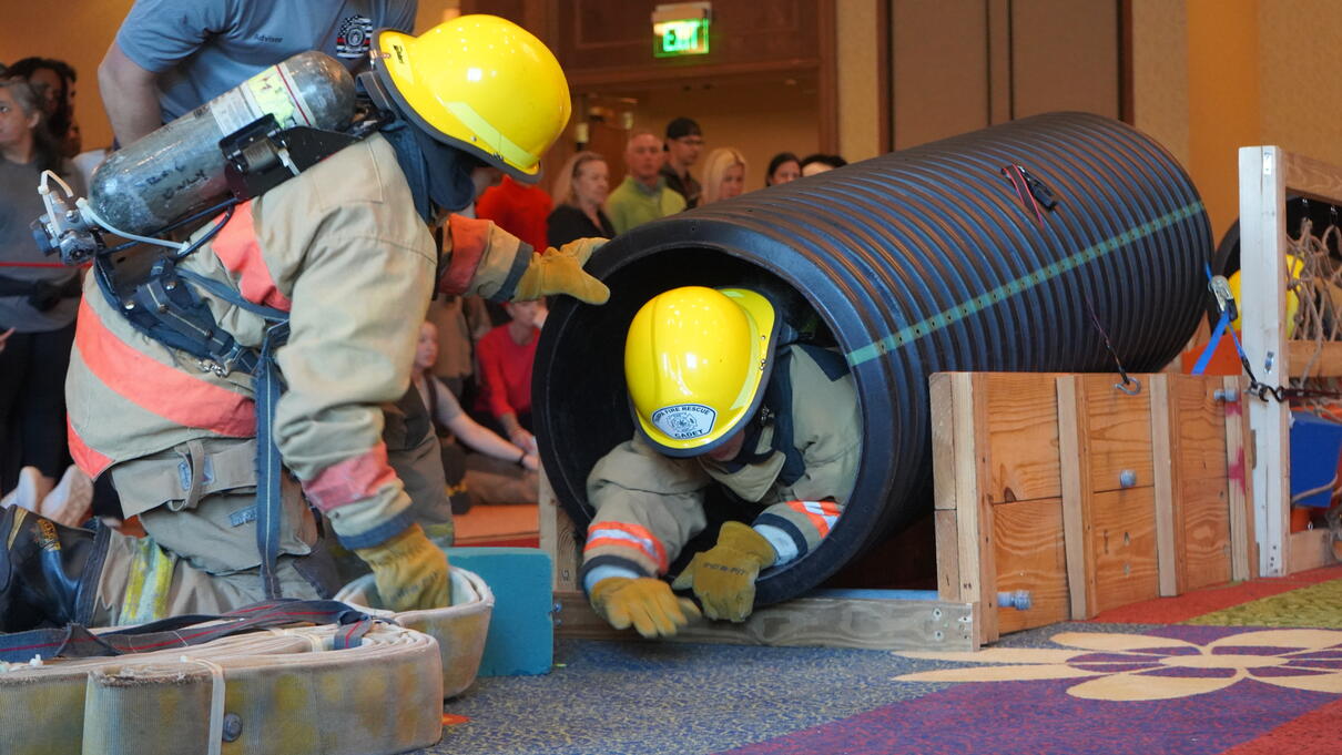 Two firefighters in full gear crawl through a black plastic tunnel during a training exercise.