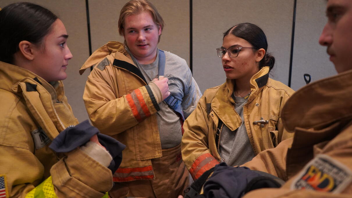 Four firefighters in uniform stand together, talking in a training or meeting setting.