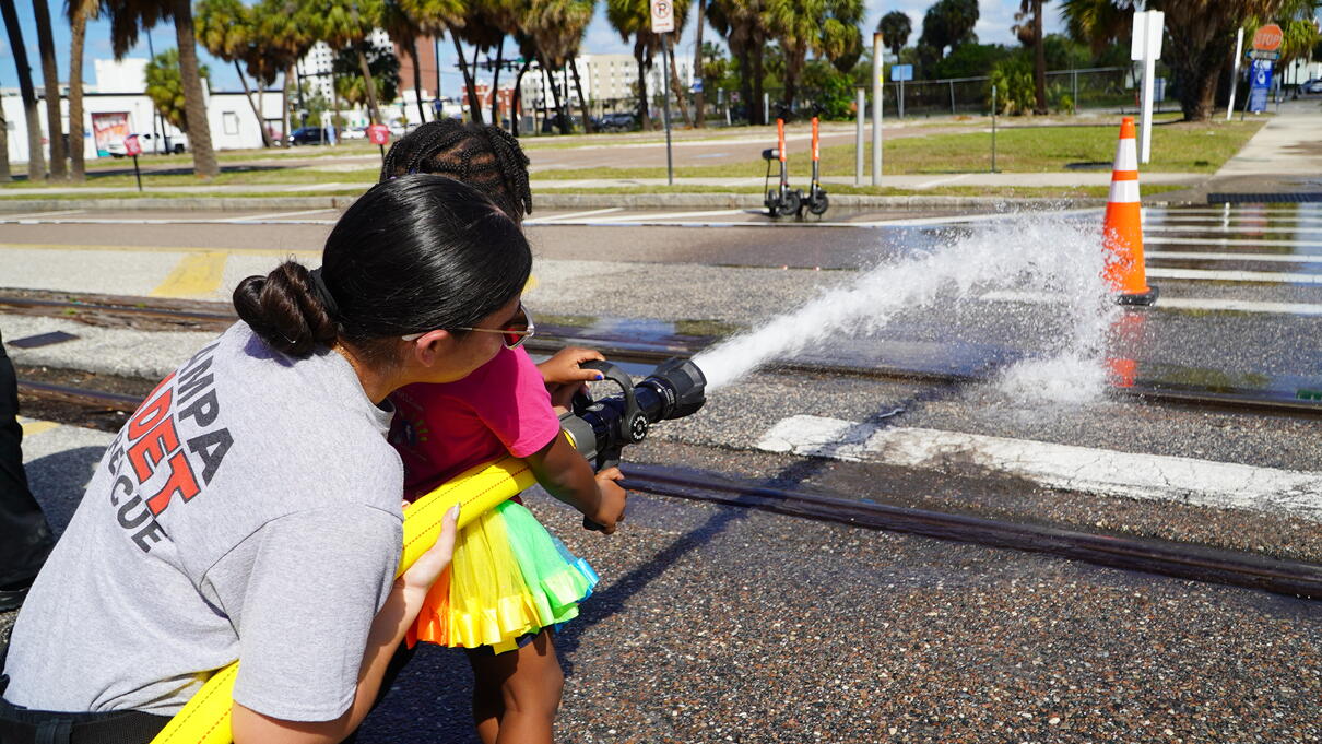 Firefighter helping young girl use hose to spray water during outdoor demonstration.