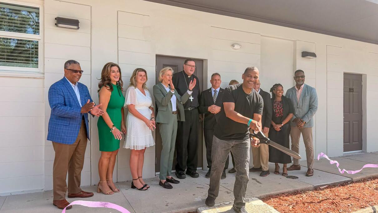 Man cutting pink ribbon at building opening ceremony with group of people applauding outside.