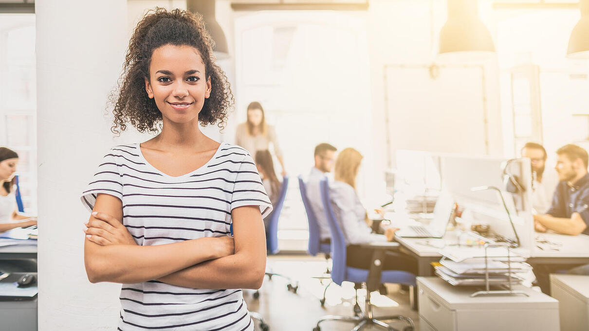 Young confident woman smiling in a busy open office setting.