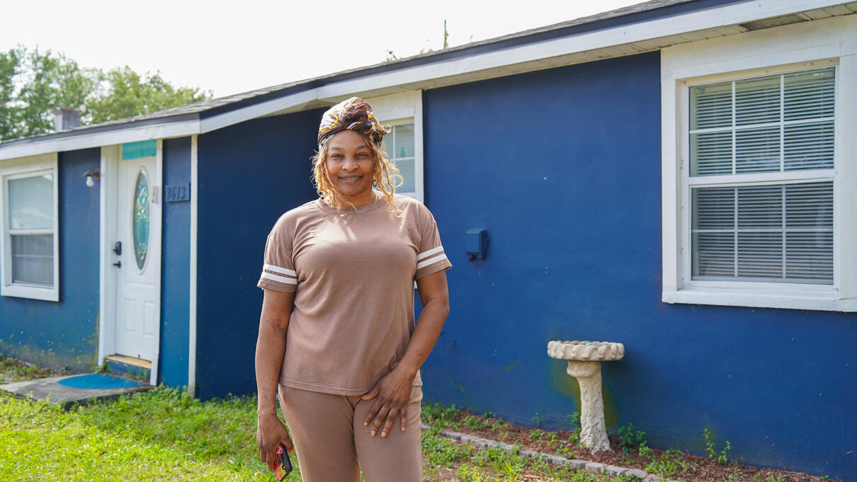 Woman smiling outside a blue house with white trim on a sunny day.