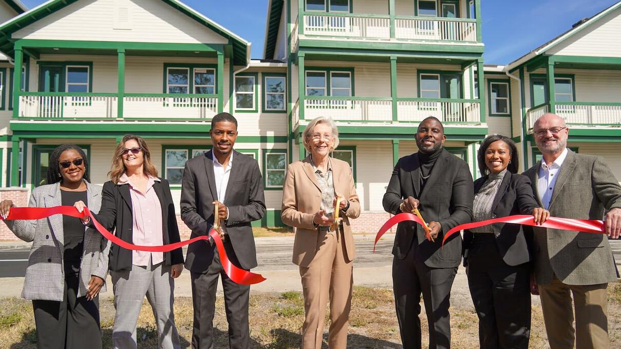 Group at a ribbon-cutting ceremony in front of green and white buildings.