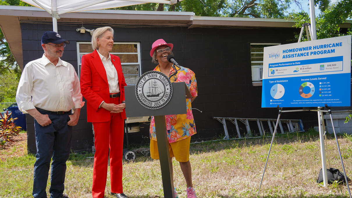 Three people stand outdoors near a signboard for a hurricane preparedness program.
