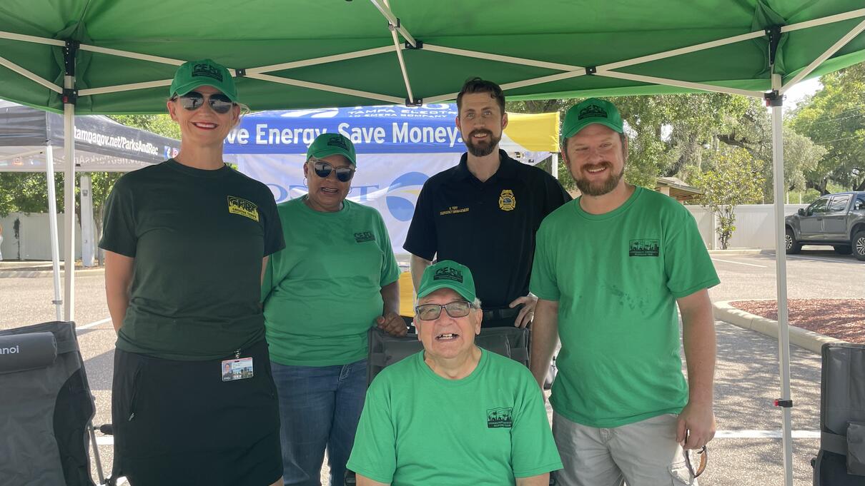 Group under a green canopy at an outdoor event, some wearing matching green shirts and caps.