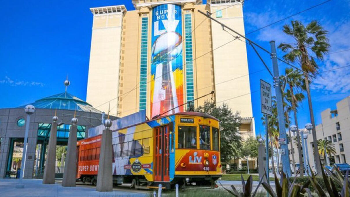 A bright streetcar passes in front of a tall building with a large Super Bowl LV banner.